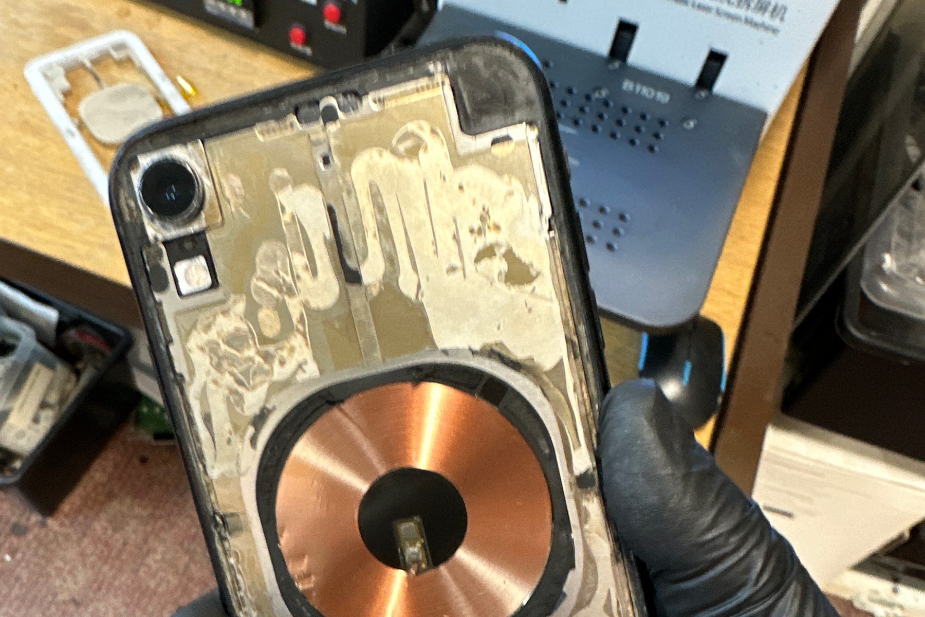 Opened smartphone held in a gloved hand, showing its internal charging coil and rear chassis on the repair bench